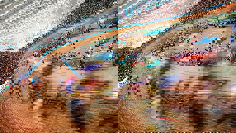 A wide-angle, blurred shot of a cycling race inside a velodrome. Cyclists are racing around a large, wooden track. The stands are filled with spectators.