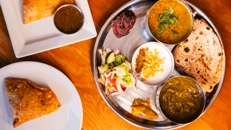 Traditional Indian thali with chapati, curries, salad, chutney, pickles, plus separate plate with samosas and dipping sauce on wooden table.