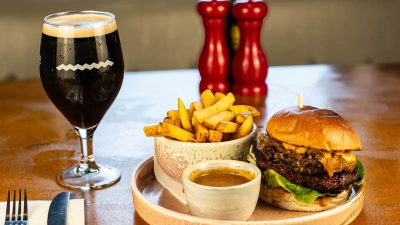 A hearty burger with melted cheese and lettuce, a bowl of chips, gravy, and a dark beer in a stemmed glass on a wooden table.