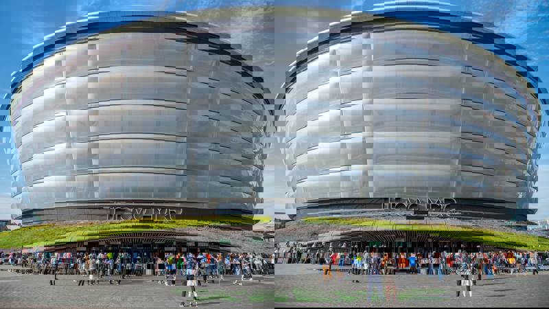 Daytime view of The OVO Hydro arena with a queue of people waiting outside under a blue sky.