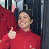 A young woman in a red fleece jacket with a logo smiles, giving a thumbs up sign on what appears to be a bus.