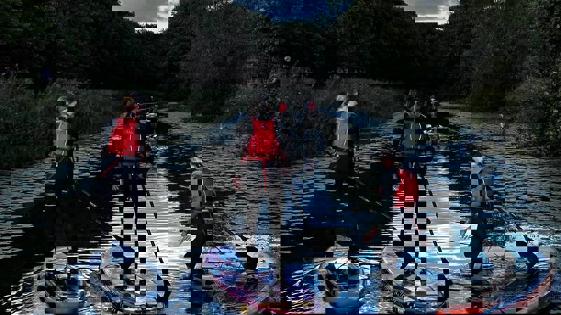 A group of people in wetsuits and red lifejackets paddleboarding along a calm tree-lined canal in Glasgow at dusk.