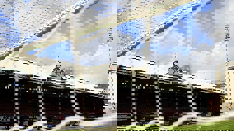 The stadium stands of a football ground are seen through the white netting of a goal. The main stand has a white roof with a triangular logo.