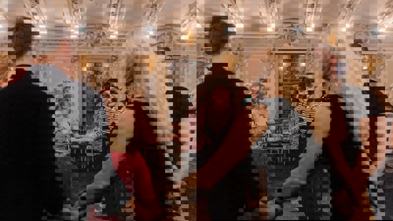 People dance in a circle at a ceilidh, a traditional Gaelic social gathering, in a room with ornate ceilings and chandeliers.