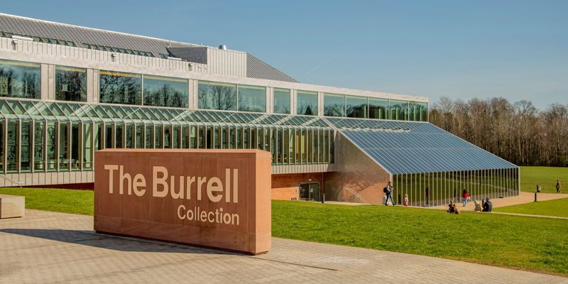 The modern, glassy Burrell Collection building is shown on a sunny day with people walking on the surrounding lawn. A large stone sign with the name of the building is in the foreground.