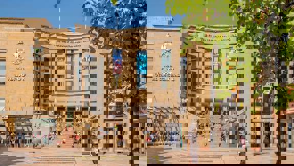 Daytime exterior of Glasgow Royal Concert Hall with people sitting on the front steps and banners promoting upcoming concerts.