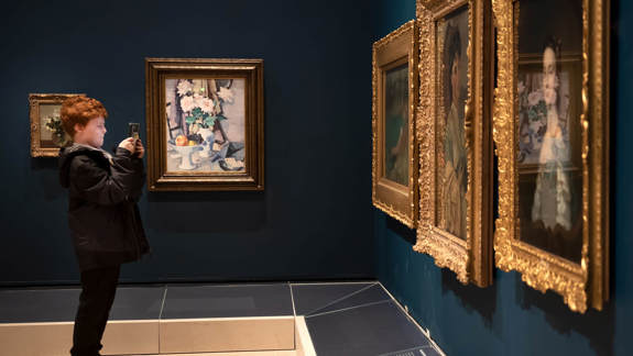 A young person uses a smartphone to take a photo of a floral still-life painting in a gallery filled with ornate gold-framed portraits.