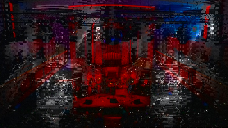 An audience watches a band play a concert in the Saint Luke's Glasgow music venue, featuring a balcony and disco ball.