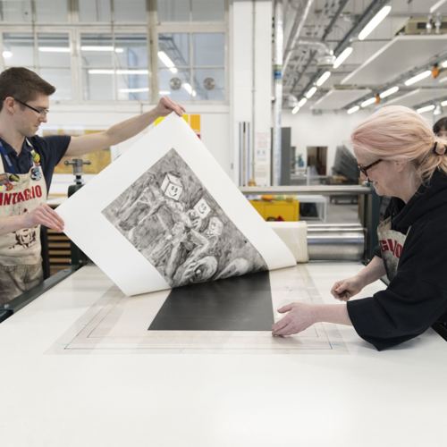 Two people in a printmaking studio carefully examine a large black-and-white print. The room is bright, organized, and filled with artistic tools.