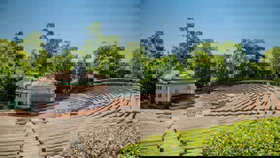 An empty outdoor amphitheatre with a small stage and tiered seating surrounded by green trees on a sunny day.