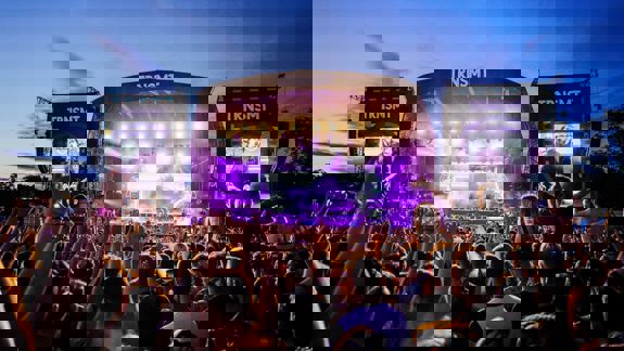 Large outdoor crowd at the TRNSMT Festival in Glasgow at dusk, with a brightly lit main stage and large video screens showing performers.