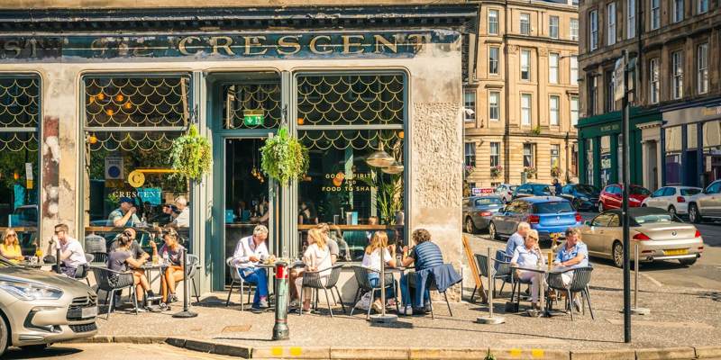 People sit at outdoor tables on a sunny day in front of a cafe with large windows and hanging plants.