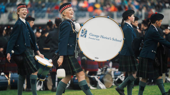 Smiling members of a marching band in traditional Scottish attire perform with drums and bagpipes at an outdoor event, with crowds in stadium seating visible in background.