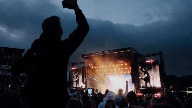 A dark shot of a crowd from behind watching a stage at a music festival. One person in the crowd stands out, as they are sitting on another person’s shoulders.