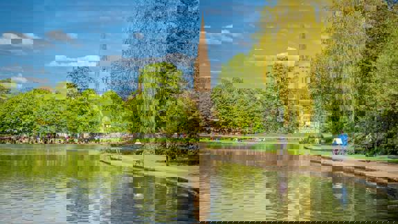 A large pond in a park with a tall church steeple in the background, with people walking and cycling on a path.