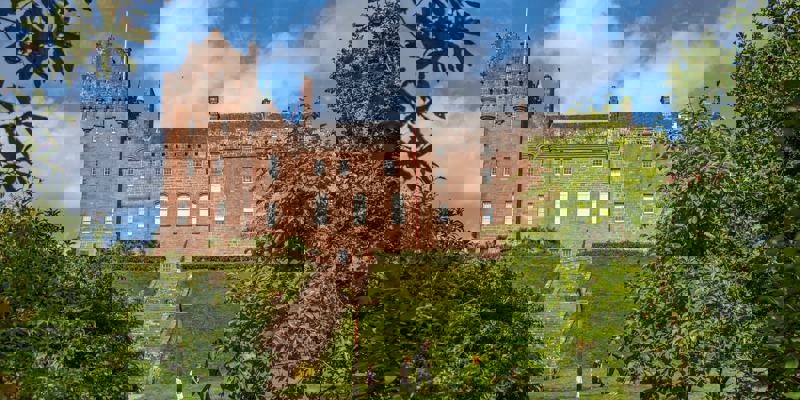 The grand red sandstone facade of Brodick Castle, featuring historic battlements and turrets, set behind lush green trees and a sloping lawn with a stone staircase.
