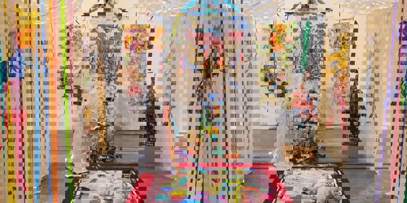  An art installation with dolls, colourful textile hangings, and figures, including one on a red rug. A figure in a white dress stands beside it.