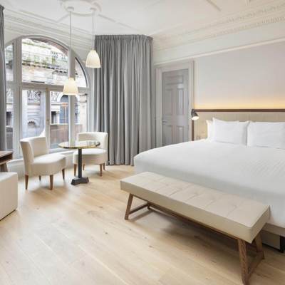 Stylish guest room at AC Hotel Glasgow with a large bed, modern lighting, light wooden floors, and a seating area beside tall arched windows overlooking the city.
