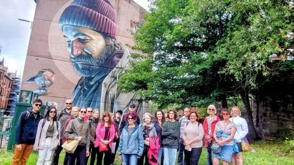 A group of people standing in front of the iconic "St Mungo" mural by Smug on High Street, Glasgow. The large-scale artwork depicts a modern-day man in a beanie with a robin.