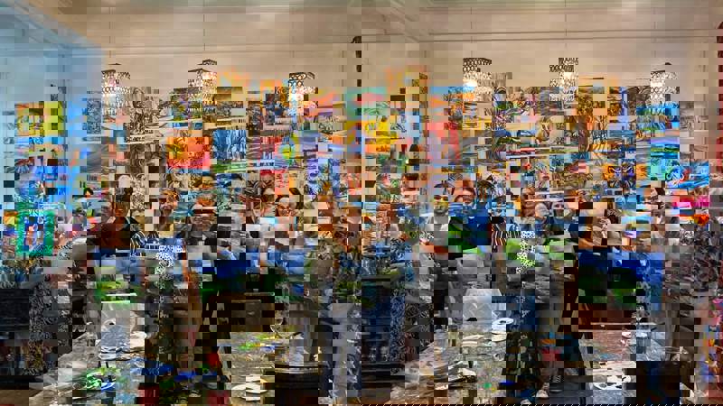 A group of people holding up their paintings after a painting class, with many other artworks on the wall behind them.