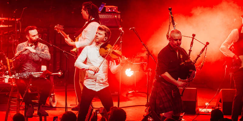 A band performs on a stage under red lighting. The musicians are playing traditional instruments, including a fiddle, a woodwind instrument, and bagpipes.