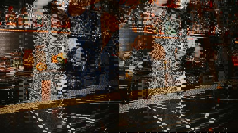 A bartender in a patterned shirt strains a cocktail into a glass on a black marble bar, with shelves of bottles behind him.