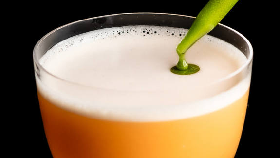 A bright orange cocktail with a frothy white head is being decorated with a dark green liquid from a pipette against a black background.