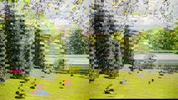 People are relaxing on a sunny, grassy hill in front of the glass Kibble Palace conservatory at the Glasgow Botanic Gardens.